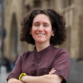 Dr. Giuditta Annibaldis: eine lächelnde Forscherin mit dunklen Locken vor einer hellbraunen Häuserfassade. Sie trägt ein weinrotes Hemd und verschränkt die Arme vor dem Körper.