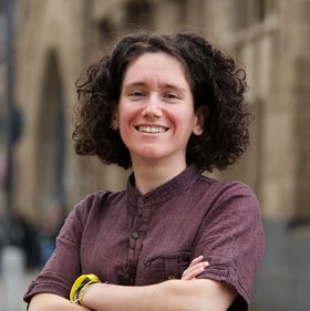 Dr. Giuditta Annibaldis Dr. Giuditta Annibaldis: eine lächelnde Forscherin mit dunklen Locken vor einer hellbraunen Häuserfassade. Sie trägt ein weinrotes Hemd und verschränkt die Arme vor dem Körper.