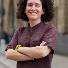 Dr Giuditta Annibaldis: a smiling researcher with dark curls in front of a light brown house façade. She wears a wine-red shirt and crosses her arms in front of her body.