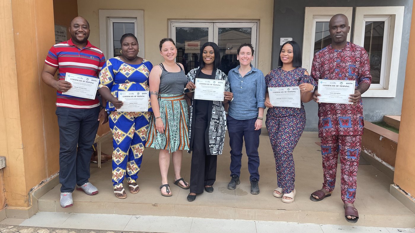 Sequencing lab staff from ISTH together with BNITM trainers at the end of the 2-weeks metagenomics sequencing training at ISTH. February 2024, Irrua, Nigeria. Five Nigerian laboratory staff and two BNITM trainers in colourful clothes are standing in the entrance of a building. They are proudly holding up their training certificates.