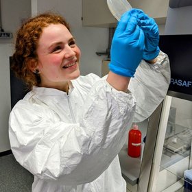 Anna-Lena Schmitz Anna-Lena Schmitz, a researcher in a lab coat holds a white object, symbolizing scientific inquiry and innovation in a laboratory setting.