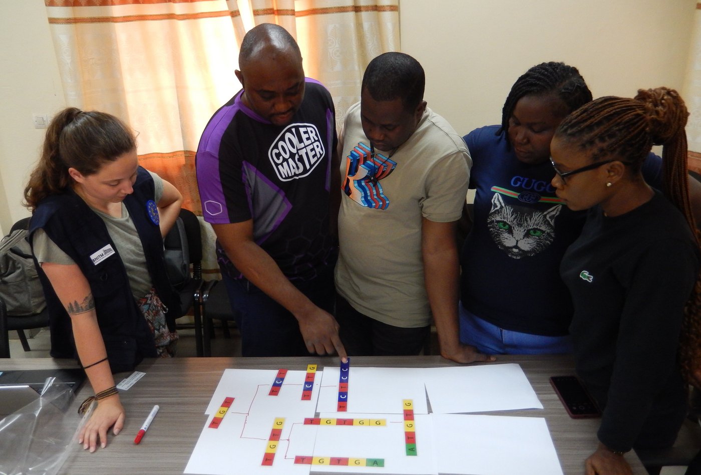 Four sequencing laboratory staff from ISTH, Nigeria, with a BNITM trainer during an introductory training on phylogenetics analysis at CRV-LFHVG, Conakry, Guinea. Das Foto zeigt vier Wissenschafter*innen mit eine Trainerin. Sie stehen vor einem zugezogenen beige-orange gemusteren Vorhang um einen Tisch und schauen bunte Buchstabenfolgen als Veranschaulichung für die durchgeführte Analyse an.