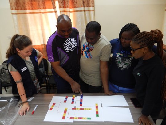 Das Foto zeigt vier Wissenschafter*innen mit eine Trainerin. Sie stehen vor einem zugezogenen beige-orange gemusteren Vorhang um einen Tisch und schauen bunte Buchstabenfolgen als Veranschaulichung für die durchgeführte Analyse an.