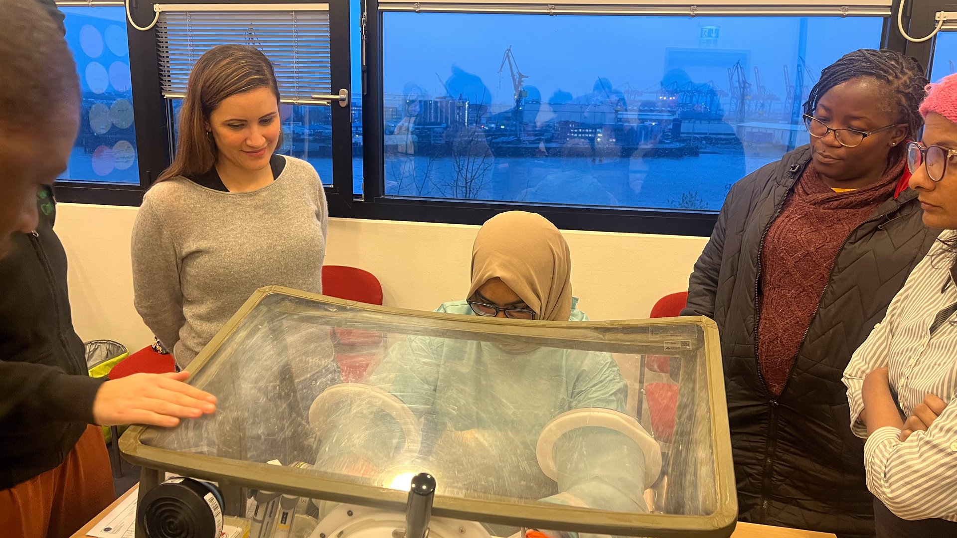 Das Foto zeigt mehrere Wissenschaftler:innen um eine Glove box herum stehen. Eine von ihnen arbeitet gerade damit.