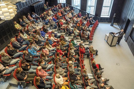 Zu sehen ist ein Hörsaal aus der Vogelperspektive. Fast alle Plätze der Sitzreihen sind besetzt und die Anwesendem lauschen einem Dozenten der vor ihnen am Pult steht.
