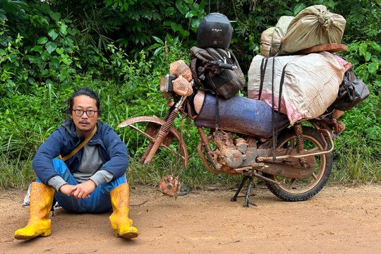The photograph shows a young man sitting on the ground next to a fully loaded motorbike.