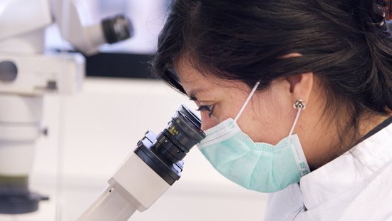 Pictured in profile is a researcher in laboratory clothing and a mouth guard. She is looking with her eyes into a microscope, which we can easily see from the side.