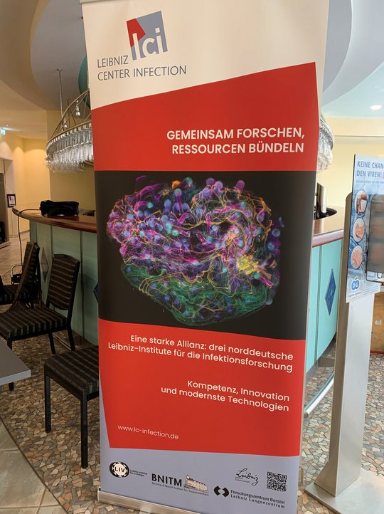 LCI Retreat Rotes Roll-up mit dem Logo des Leibniz Center Infection, einem farbigen wissenschaftlichen Bild und Text zu gemeinsamer Infektionsforschung.