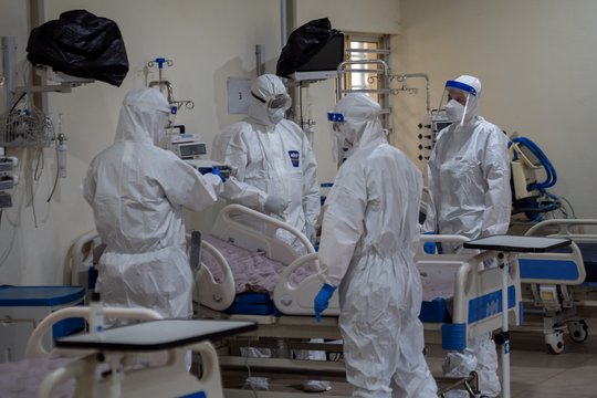 MEVIN 2.0 Das Bild zeigt vier Personen in weißen Schutzanzügen mit Mundschutz, Gesichtsschutz und blauen Handschuhen um ein leeres Krankenhausbett herum stehen.