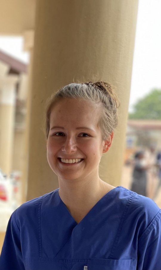 Das Bild zeigt eine Frau, die in die Kamera lächelt. Sie trägt einen blauen Krankenhauskittel.