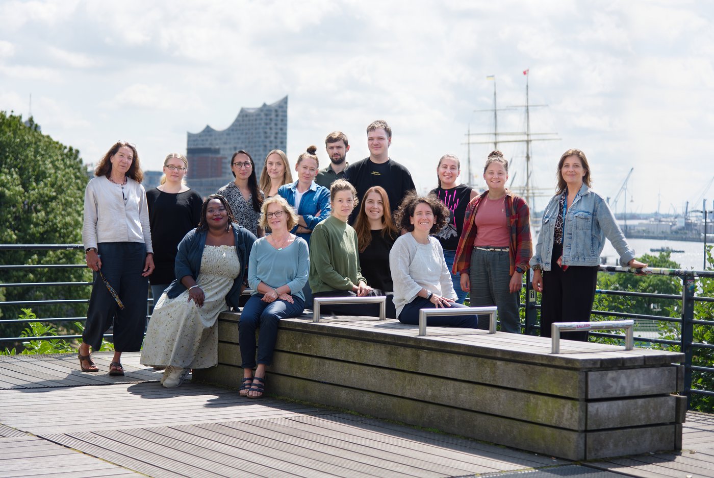The picture shows a team of 15 sitting on a bench or standing behind it on a sunny day. In the background you can see the building of the Hamburg Elbphilharmonie.