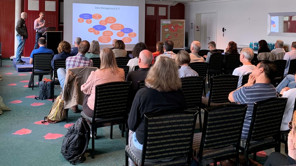 LCI Retreat Ein Referent steht in einem Konferenzraum vor einem Publikum und präsentiert eine Folie mit dem Titel „Datenmanagement & IT“, auf der ein Flussdiagramm aus orangefarbenen und blauen Kreisen zu sehen ist.
