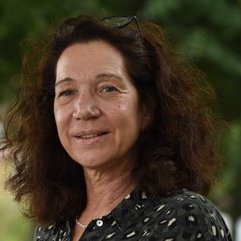 Beate Becker-Ziaja: a woman with shoulder-length, wavy brown hair into which glasses are tucked smiles at the camera. Leaves can be seen blurred in the background.