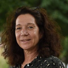 Beate Becker-Ziaja: a woman with shoulder-length, wavy brown hair into which glasses are tucked smiles at the camera. Leaves can be seen blurred in the background.