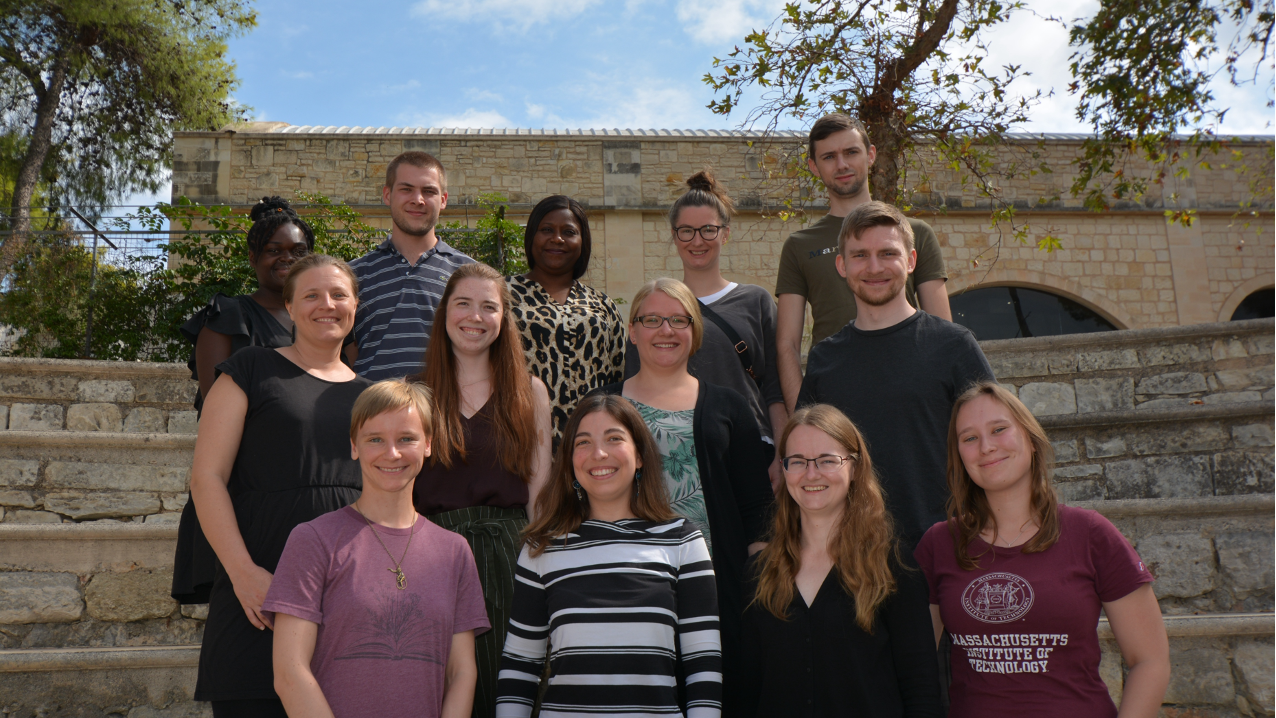 The members of the Junior Research Group Oestereich stand next to each other with smiling faces for a group picture. Some trees and a building are visible in the background.