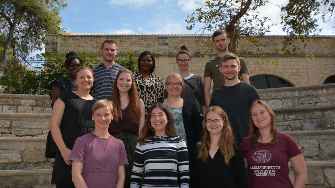 Research Group Oestereich. The members of the Junior Research Group Oestereich stand next to each other with smiling faces for a group picture. Some trees and a building are visible in the background.