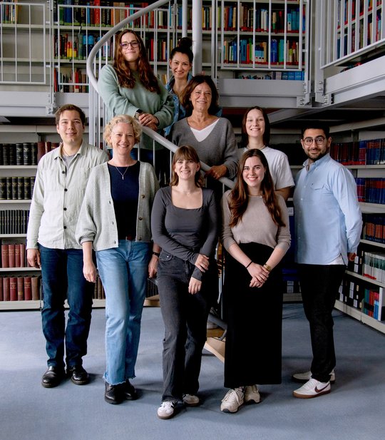 The working group with 9 members is standing at spiral stairs in the library of the Bernhard Nocht Institute. the group is posing infront of  the staircase, creating a beautiful overall picture. Everyone smiles into the camera.