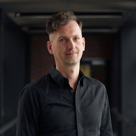 Dr. Ralf Krumkamp: ein Forscher mit kurzen, braun-grauen Haaren, trägt ein schwarzes langes Hemd und eine Jeans.
