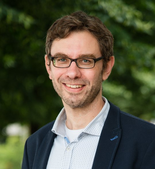 Jan Strauss Auf dem Foto ist ein junger Mann mit kurzen braunen Haaren und einer Brille zu sehen. Er trägt ein helles Hemd und ein schwarzes Jacket.