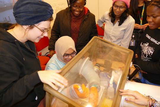 The foto shows 5 women standing around a glove box. One of them is training to handle materials in the glove box.