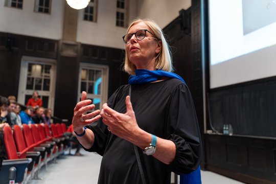 Auf dem Foto ist Petra Emmerich zu sehen, die im Hörsaal einen Vortrag hält. Rechts im Hintergrund ist die Leinwand mit Folie zu sehen, links in Hintergrund ein paar Sitzreihen mit roten Stühlen.