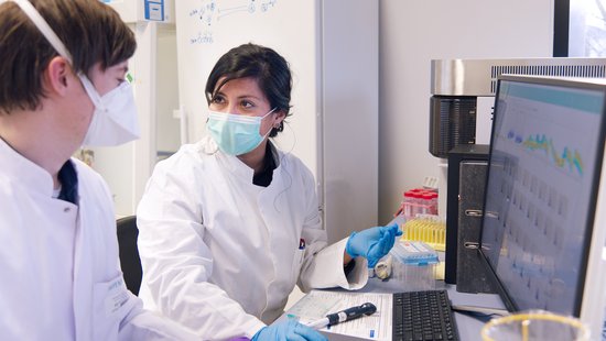 In the lab The photo shows two researchers, a woman and a man, wearing face masks and lab coats, sitting in front of a screen and talking.