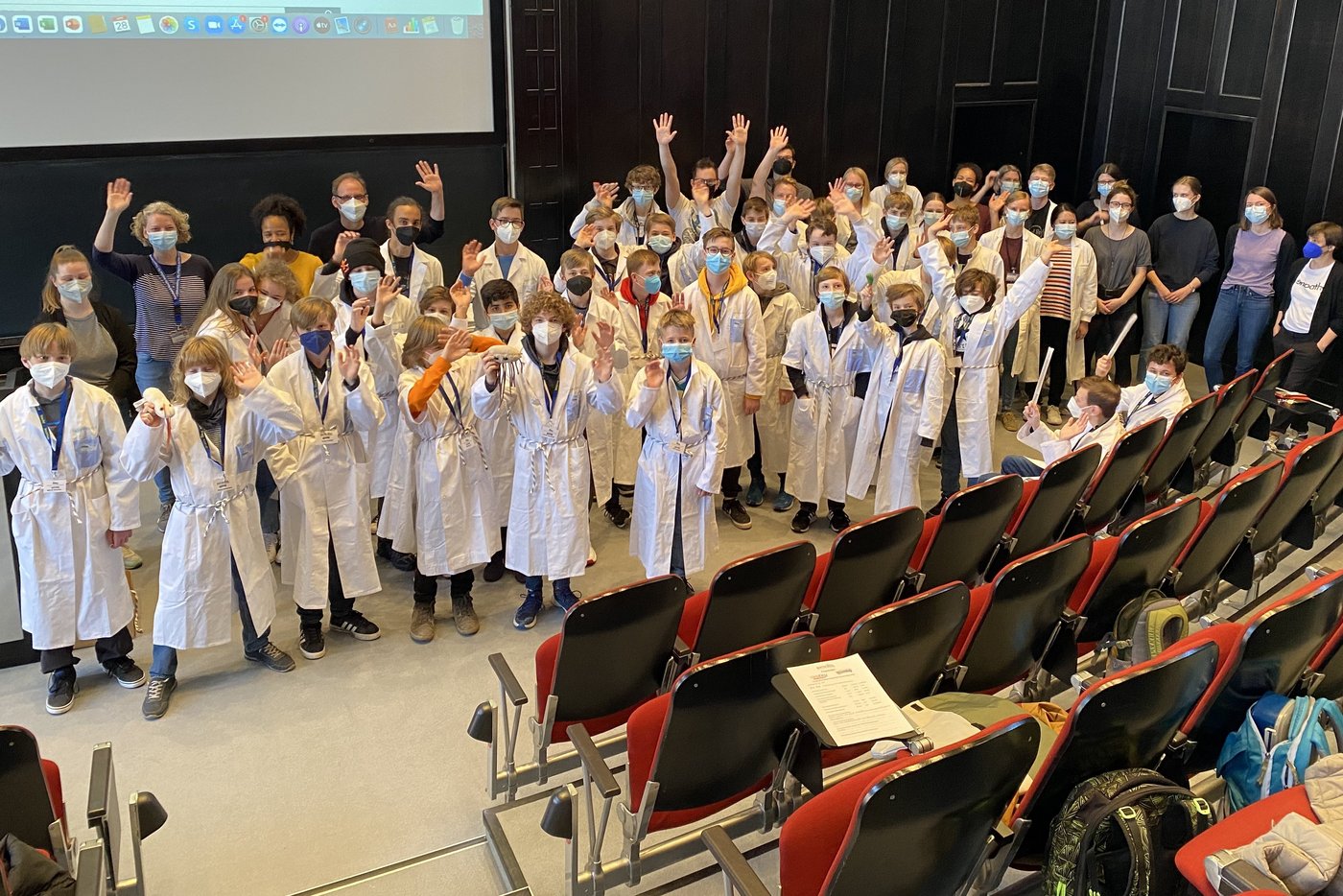 Etwa 30 Jugendliche stehen mit Laborkitteln bekleidet mit einigen Forscher:innen im Hörsaal vor der Tafel und winken.