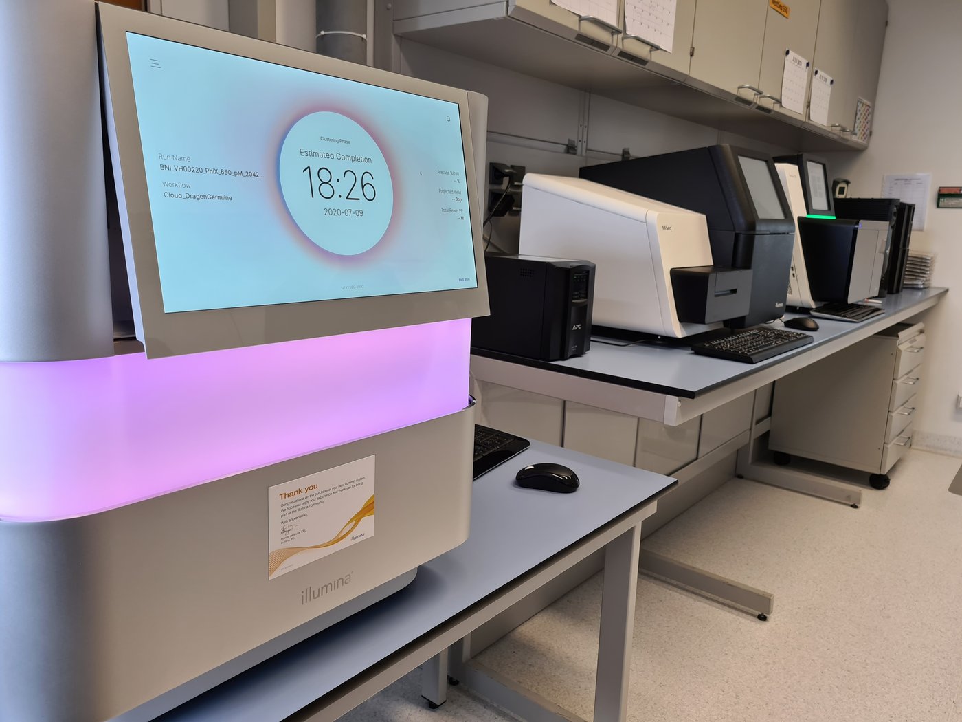 The Next-Generation Sequencing (NGS) unit of the BNITM can be seen. In the foreground is a large white box on a table. In the middle runs a wide luminous strip that glows pink-purple. At the top, you can see a large screen with the text estimated completion 18:26. In the background, there is more laboratory equipment on tables.