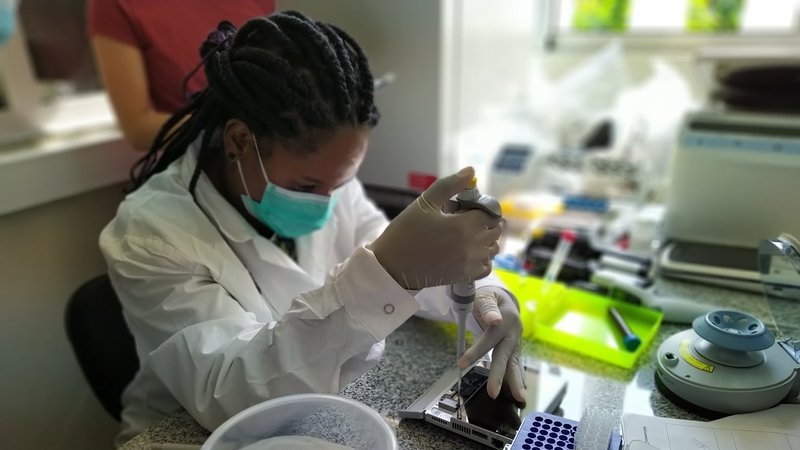 Eine Wissenschaftlerin übt konzentriert ein labordiagnostisches Verfahren an einem Arbeitstisch. Um sie herum sind diverses Equipment für die Labordiagnostik und Dokumente verteilt.