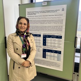 Dr. Molouk Beiromvand Eine Person steht in Mantel und Kopftuch gekleidet neben einer wissenschaftlichen Posterpräsentation zur Diagnose von Strongyloidiasis im Iran in einem Raum mit Fenstern.