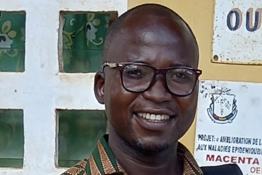 Raymond Koundouno: A smiling man wearing dark rimmed glasses. He wears a green, red and beige patterned shirt and is standing in front of a yellow wall with signs on it.