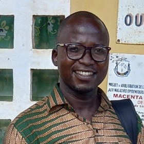 Raymond Koundouno: einen lächelnden Mann mit Brille. Er trägt ein grün-rot-beige gemustertes Hemd und steht vor einer Hauswand mit Schildern.