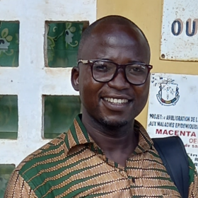 Raymond Koundouno: a smiling man with glasses. He wears a green-red-beige patterned shirt and stands in front of a house wall with signs.