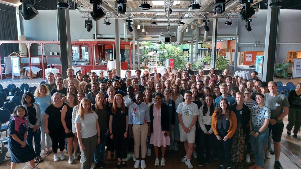 The photo shows a large group of young scientists in a factory-like hall.