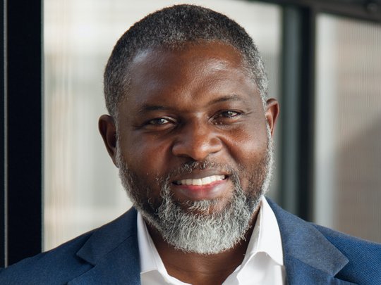 The portrait photo shows Ghanaian researcher John Amuasi. He has a slightly greying beard, is wearing a white shirt and blue jacket, and is smiling at the camera.