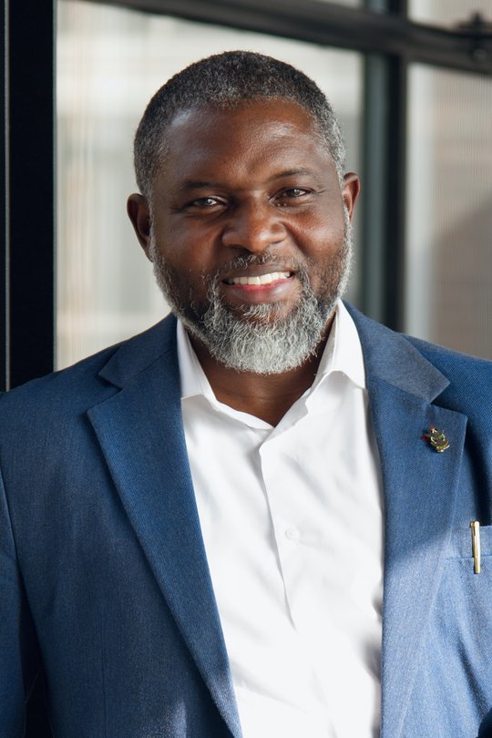 Prof. Dr. John Amuasi Das Porträt-Foto zeigt den ghanaischen Forscher John Amuasi. Er trägt einen leicht ergrauten Bart, ein weißes Hemd und ein blaues Sakko und lächelt in die Kamera.