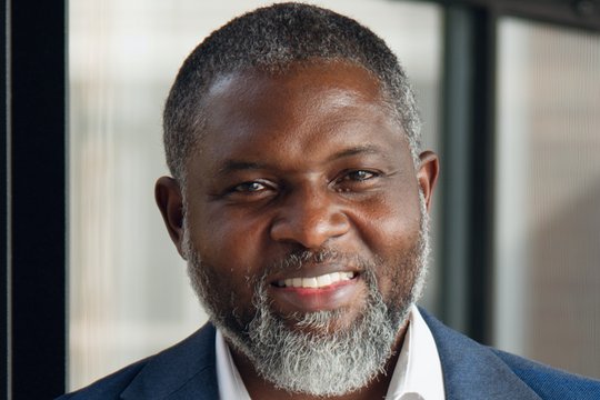 Das Porträt-Foto zeigt den ghanaischen Forscher John Amuasi. Er trägt einen leicht ergrauten Bart, ein weißes Hemd und ein blaues Sakko und lächelt in die Kamera.