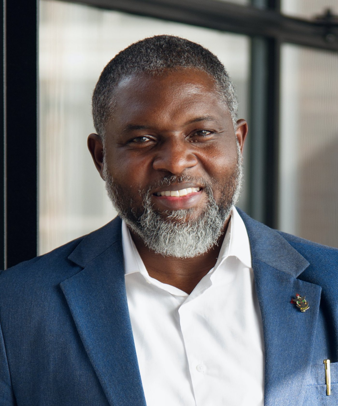 Prof. Dr. John Amuasi Das Porträt-Foto zeigt den ghanaischen Forscher John Amuasi. Er trägt einen leicht ergrauten Bart, ein weißes Hemd und ein blaues Sakko und lächelt in die Kamera.