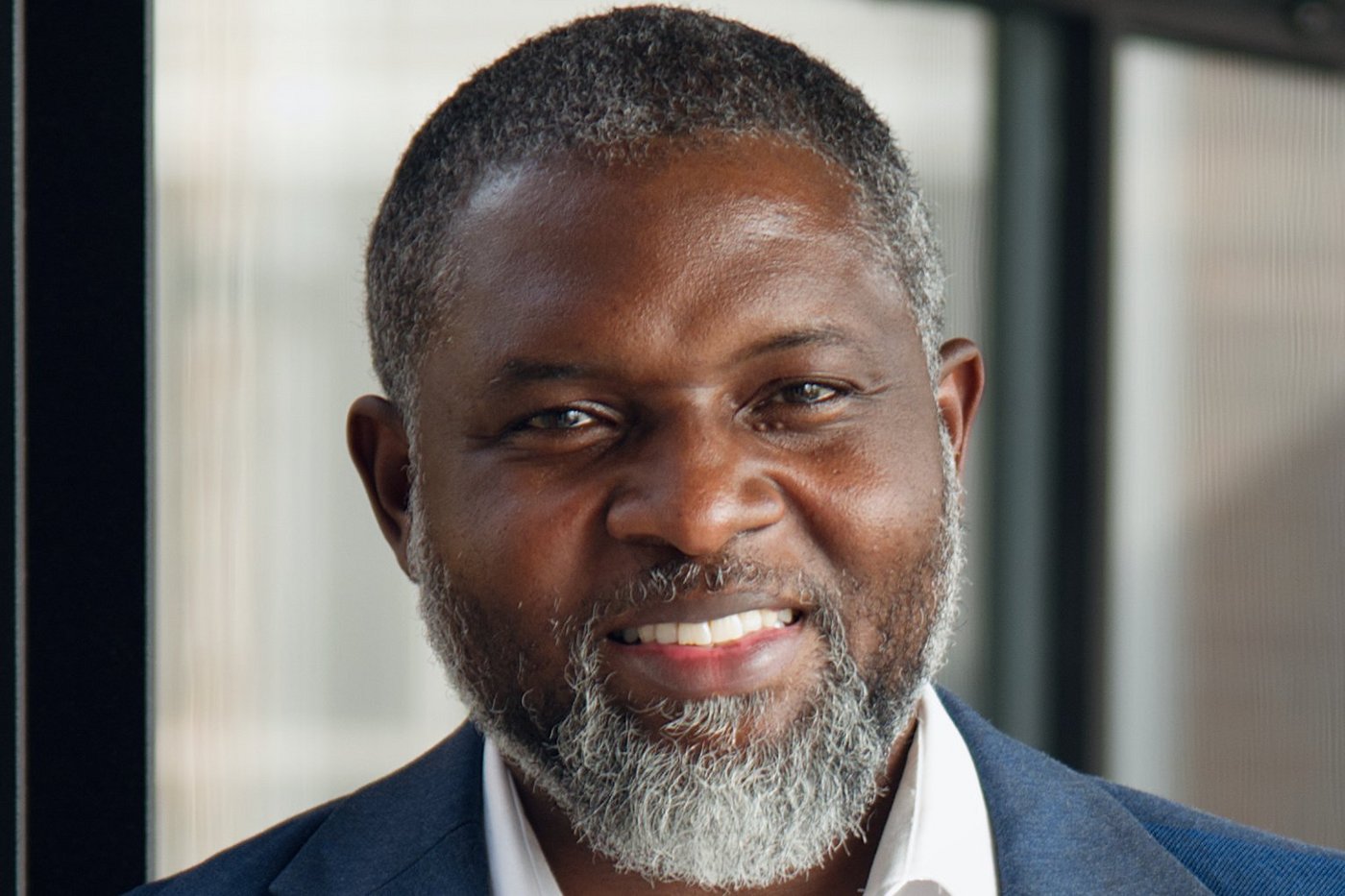 The portrait photo shows the Ghanaian researcher John Amuasi. He wears a slightly greying beard, a white shirt and a blue jacket and smiles at the camera.
