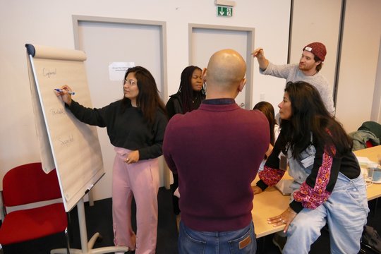 The foto shows 5 persons doing group work around a table. One of them writes on a flip chart.