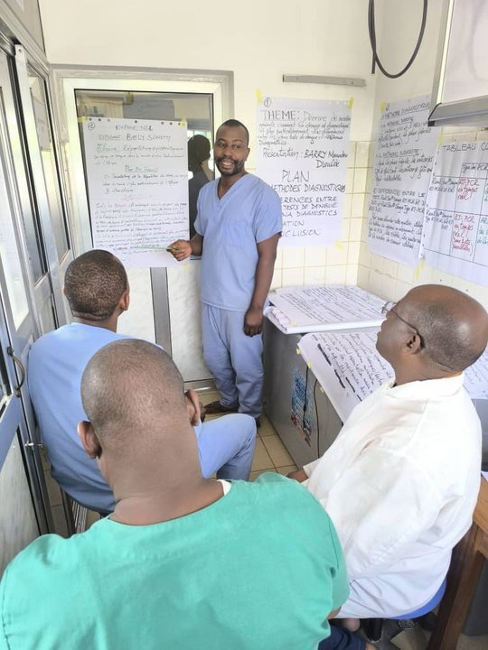 Three people are sitting in the laboratory while another laboratory member is presenting a poster at the front. Several posters are hanging on the back wall.