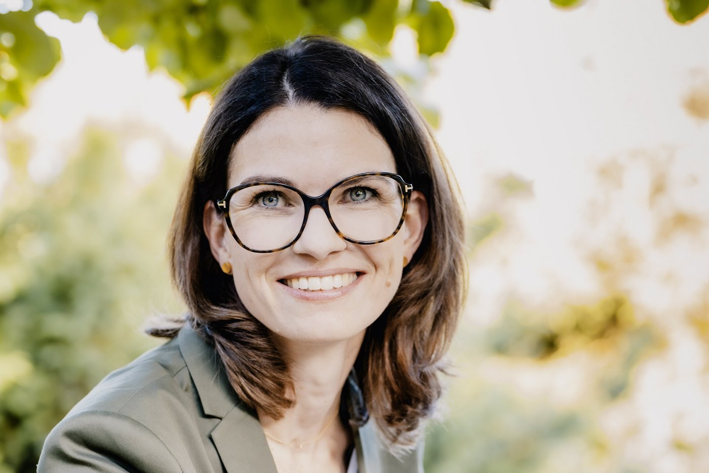 Prof. Dr. Cornelia Betsch: eine Forscherin mit braunen, schulterlangen Haaren, Brille und einem grauen Blazer über einem hellen Oberteil. Sie sitzt im Freien, im Hintergrund sind Blätter zu sehen.