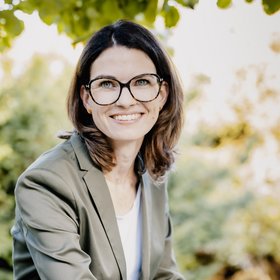 Prof. Dr. Cornelia Betsch: eine Forscherin, die braunes, schulterlanges Haar, eine Brille und einen grauen Blazer über einem hellen Oberteil trägt. Sie sitzt draußen, im Hintergrund sind Blätter zu sehen.