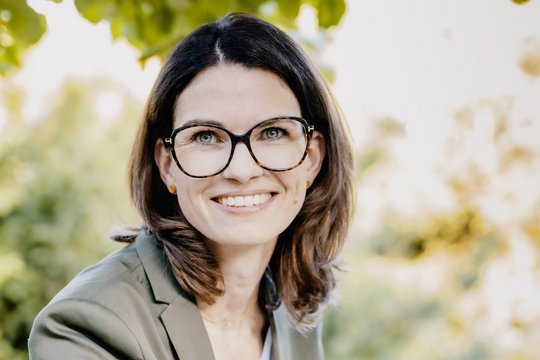 Prof. Dr Cornelia Betsch: a researcher wearing brown, shoulder-length hair, glasses and a grey blazer over a light-coloured top. She is sitting outside, leaves can be seen in the background.
