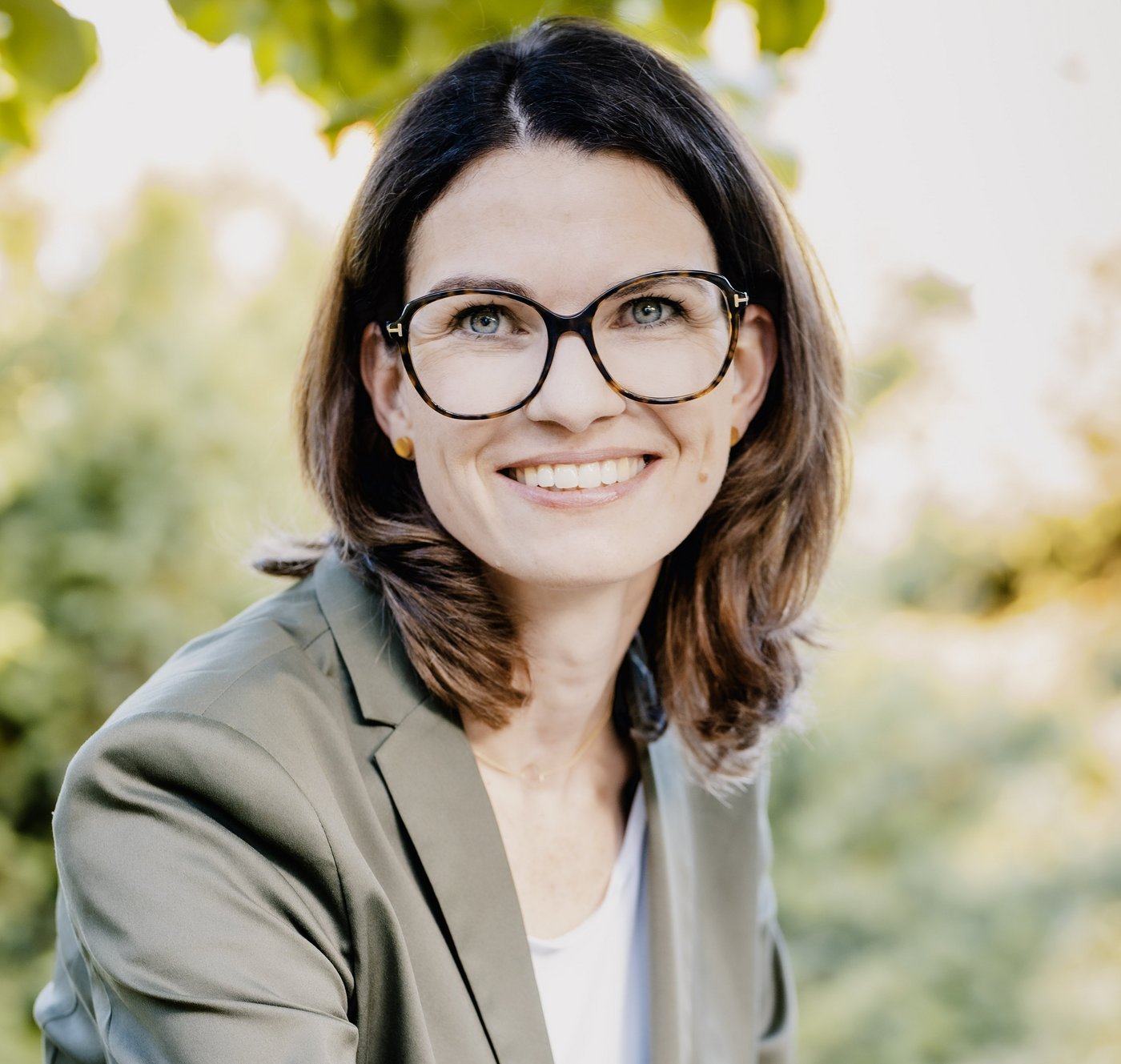 Prof. Dr. Cornelia Betsch Prof. Dr. Cornelia Betsch: eine Forscherin mit braunen, schulterlangen Haaren, Brille und einem grauen Blazer über einem hellen Oberteil. Sie sitzt im Freien, im Hintergrund sind Blätter zu sehen.