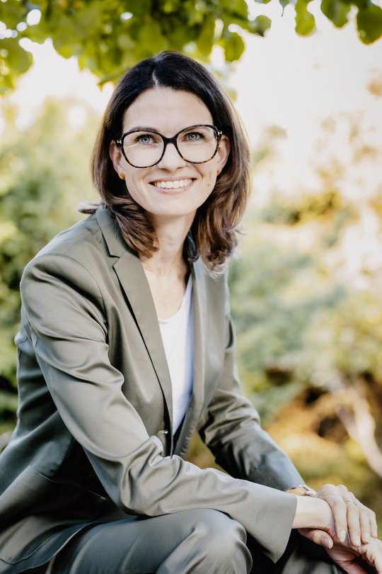 Prof. Dr. Cornelia Betsch: eine Forscherin mit braunen, schulterlangen Haaren, Brille und einem grauen Blazer über einem hellen Oberteil. Sie sitzt im Freien, im Hintergrund sind Blätter zu sehen.