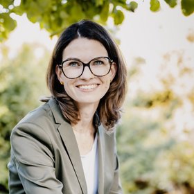 Prof. Dr. Cornelia Betsch: a researcher wearing brown, shoulder-length hair, glasses and a grey blazer over a light-coloured top. She is sitting outside, leaves can be seen in the backgrou