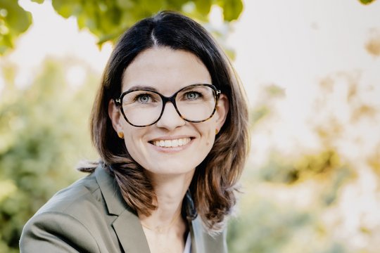 Prof. Dr. Cornelia Betsch: eine Forscherin mit braunen, schulterlangen Haaren, Brille und einem grauen Blazer über einem hellen Oberteil. Sie sitzt im Freien, im Hintergrund sind Blätter zu sehen.