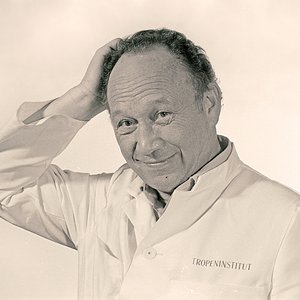 Dr. Günther Müller 1986 Schwarz-Weiß-Foto von Dr. Günther-Müller im weißen Laborkittel. Er schaut freundlich in die Kamera und kratzt sich mit der rechten Hand am Hinterkopf.
