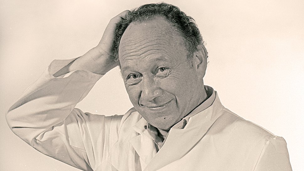 A black-and-white photograph of Dr Günther-Müller in a white lab coat. He is smiling warmly at the camera and scratching the back of his head with his right hand.